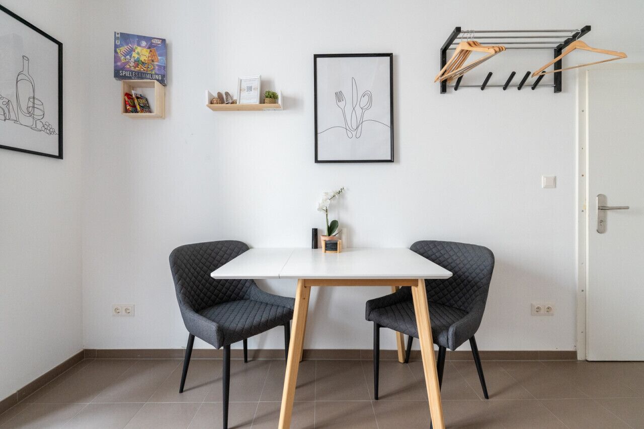 A modern room with a white table, two black chairs, a framed picture of cutlery, a small shelf with items, and a door with a towel rack.