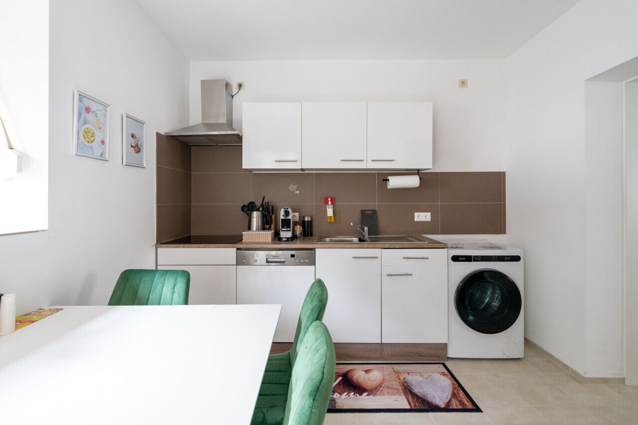 Una cucina compatta con mobili bianchi, lavastoviglie, lavatrice e zona pranzo con una sedia verde.