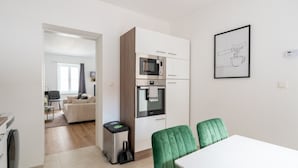 Eine moderne Küche mit Einbauofen und Mikrowelle, ein Essbereich mit weißem Tisch und grünen Stühlen sowie ein gerahmtes Bild an der Wand.