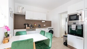 Eine moderne Küche mit weißen Schränken, einer Edelstahl-Dunstabzugshaube, einer Mikrowelle und einem Backofen. Ein Esstisch mit grünen Stühlen und einem kleinen Topf mit Pflanze steht dort.