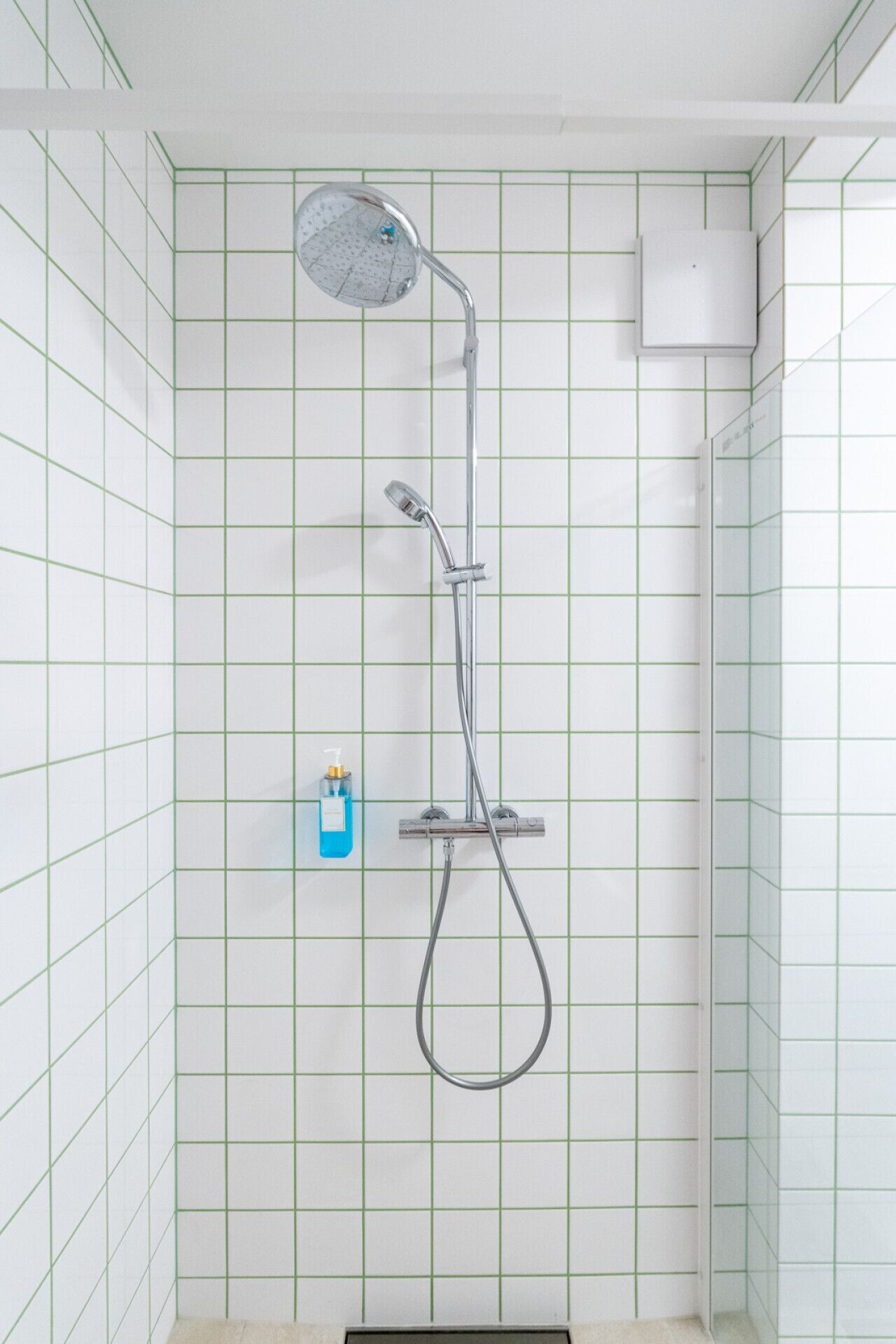 A shower with a handheld showerhead and a wall-mounted faucet, set against a tiled wall with a grid pattern.