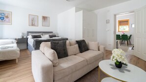 Ein modernes, helles Wohnzimmer mit einer Couch, einem Bett und einem Essbereich, der durch eine offene Tür sichtbar ist.