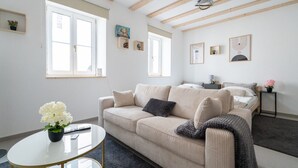 Un soggiorno moderno con un divano color beige, un tavolino da caffè e un letto sullo sfondo. Due finestre lasciano entrare luce naturale, e l'ambiente presenta un design minimalista con alcuni elementi decorativi alle pareti.