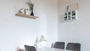 A dining area with a white table, four grey upholstered chairs, and a wooden floor. There are two shelves with plants and a whiteboard on the wall.