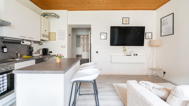 Une cuisine moderne avec un plafond en bois, un îlot central doté de tabourets de bar, un téléviseur à écran plat fixé au mur et un espace de vie minimaliste avec un canapé et une lampe.