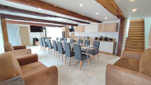 Un comedor amplio con una mesa larga dispuesta para seis personas, rodeado de sofás cómodos. Se ve una cocina con electrodomésticos modernos al fondo.