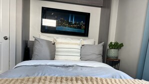 A neatly made bed with a grey duvet, a beige knitted throw, and a cityscape TV screen above. - Blueberry Bungalow with Stunning Patio & Garden (Dayton)