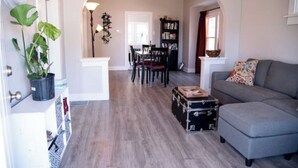 A living room with a grey sofa, a dining area with a table and chairs, and a bookshelf. - Blueberry Bungalow with Stunning Patio & Garden (Dayton)