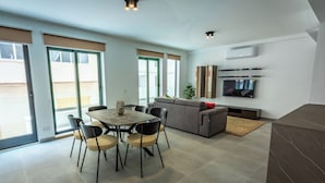 A modern living room with a dining area, a grey sofa, a flat-screen TV mounted on the wall, and a sliding glass door leading to an outdoor area.