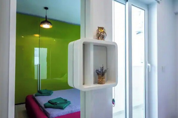 Una habitación con pared verde, un estante blanco con una decoración de lechuzas, una lámpara colgante y una cama con ropa de cama verde y rosa.