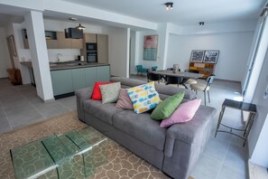 Un espacio moderno para estar con un sofá gris, cojines coloridos, una mesa de centro de vidrio, una mesa de comedor con sillas y una cocina al fondo.