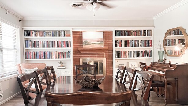 A dining room with a large wooden table, surrounded by chairs, and flanked by bookshelves filled with books. A fireplace is visible in the center, and a piano is placed against the wall. - The Cozy Home near Lancium, ACU, HSU, Hendrick (Abilene)