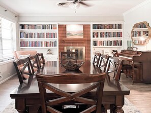 Un comedor con una gran mesa de madera, rodeada de sillas y flanqueada por estanterías llenas de libros. Se observa una chimenea en el centro, y un piano apoyado contra la pared.