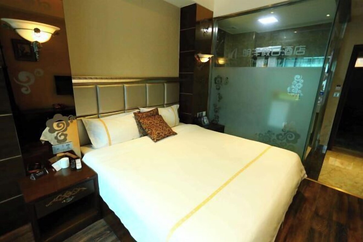 A hotel room with a large bed, a nightstand with a lamp, and a glass door leading to a bathroom.
