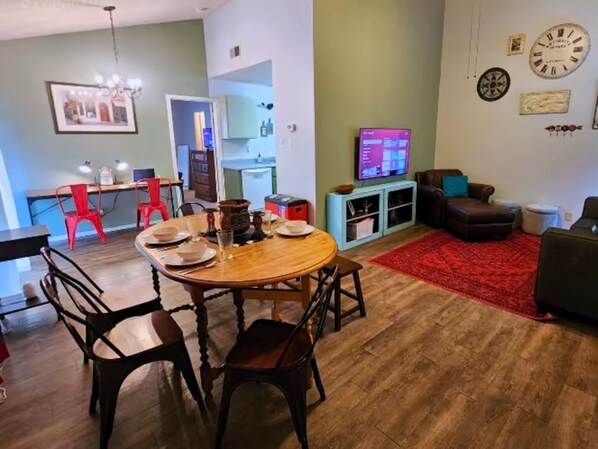 A dining area with a wooden table and chairs, a red rug, a television, and a clock on the wall.