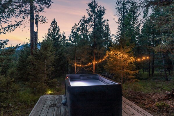 A hot tub on a wooden deck surrounded by trees, with string lights in the background.