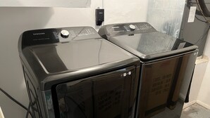 Dos lavadoras de carga frontal negras en un cuarto de servicio, con un enchufe visible y un respiradero.