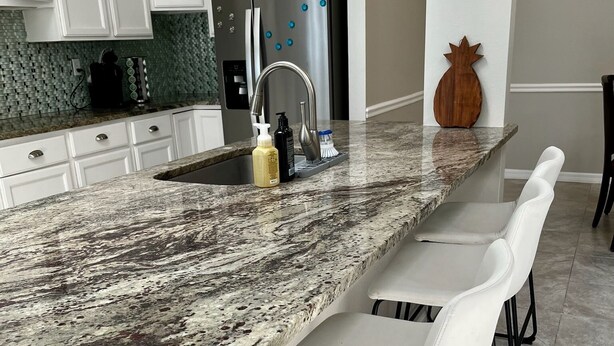 Una cocina con armarios blancos, encimera de granito, refrigerador de acero inoxidable y taburetes de bar.