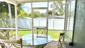 Un patio cubierto con una mesa redonda de vidrio y sillas de mimbre, con vistas a un jardín trasero con una valla blanca y follaje.