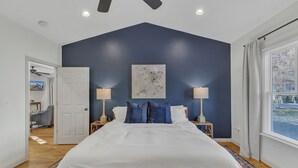 Quarto com uma cama grande, duas mesas de cabeceira com abajures, um ventilador de teto, um quadro na parede e uma janela com cortinas.
