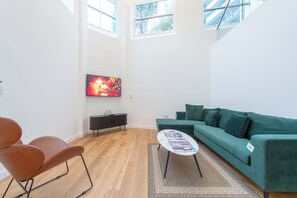 A modern living room with a green sofa, a brown leather chair, a coffee table with magazines, a TV on the wall, and a window with a view of trees. - Exquisite 3BR Duplex Apartment, Air-con& Amenities (London)