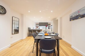 A modern dining area with a wooden floor, a dining table set for four, and a kitchen in the background. - Exquisite 3BR Duplex Apartment, Air-con& Amenities (London)