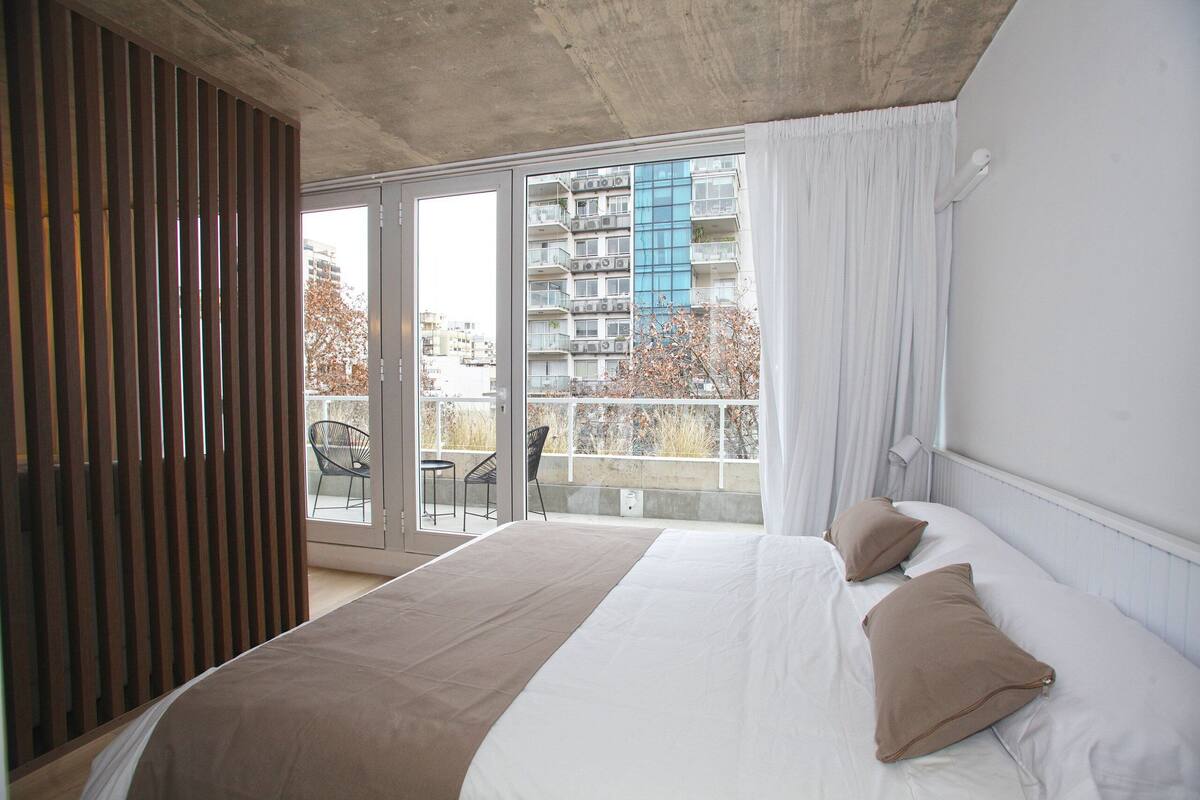Un dormitorio con una cama grande, una puerta corrediza al balcón y vistas de un edificio con varios balcones.