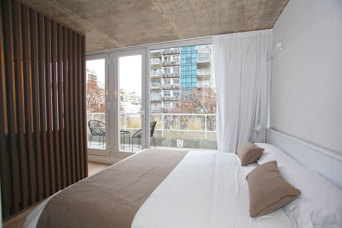 A bedroom with a large bed, a sliding glass door to a balcony, and a view of a building with multiple balconies.