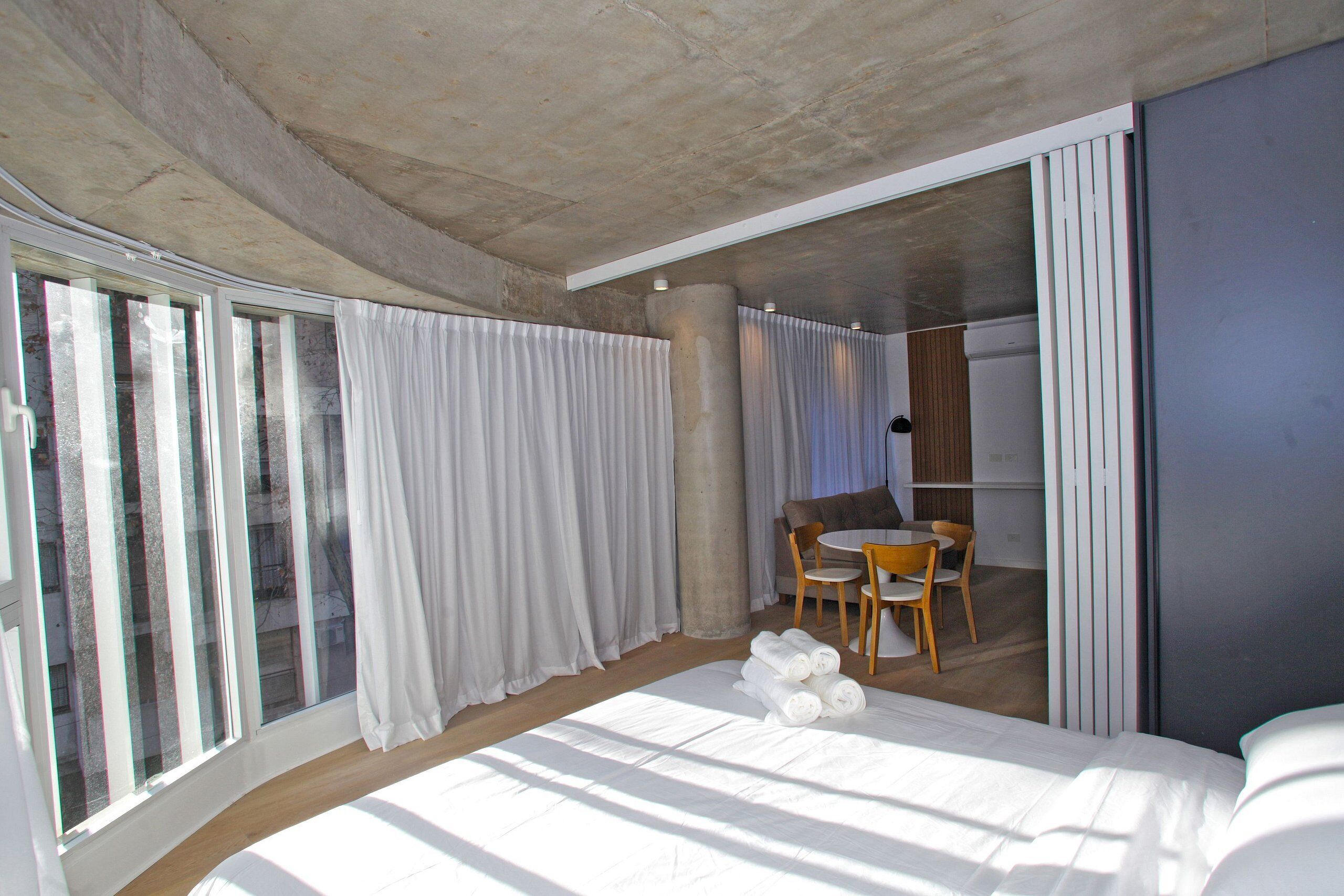 Una habitación de hotel moderna con una cama grande, un área de comedor con mesa y sillas, y un ventanal corredizo que da a un balcón.