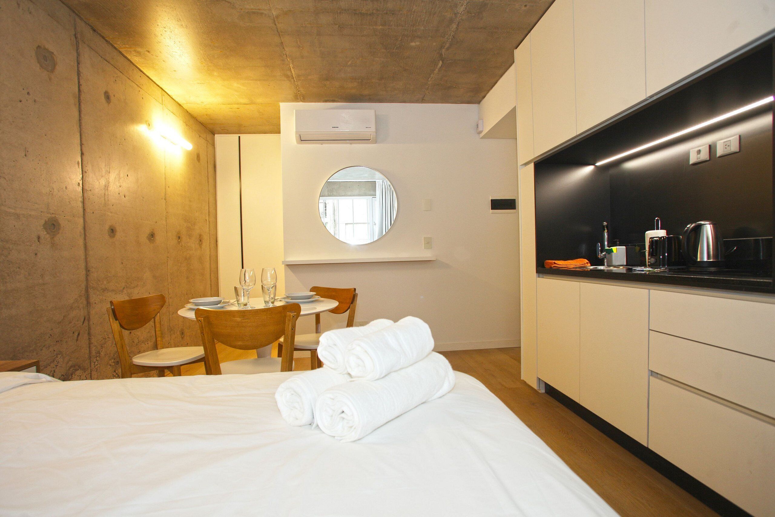 A hotel room with a bed, a round mirror, a small dining table with chairs, and a kitchenette.