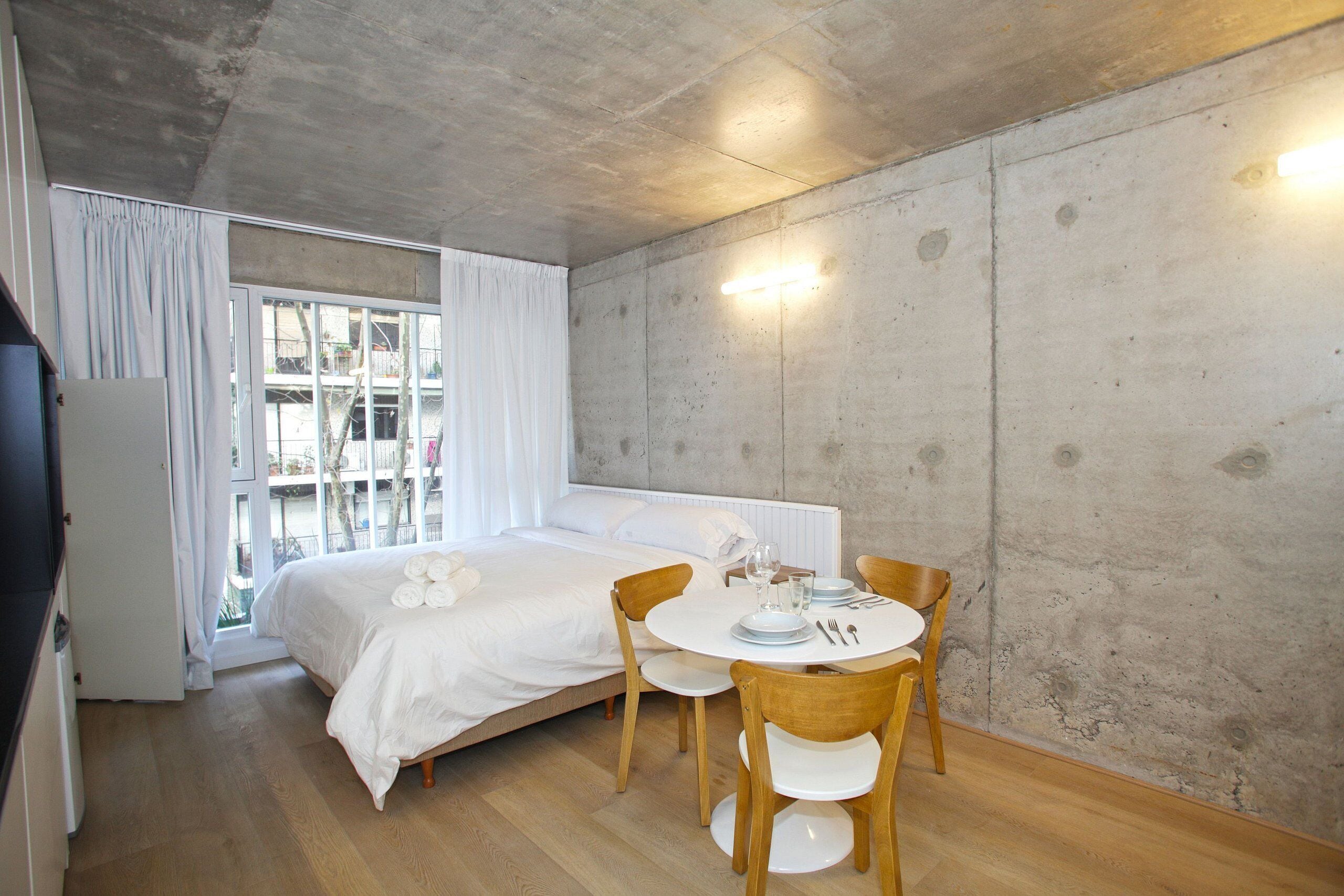 A bedroom with a bed, a dining table with chairs, a window with curtains, and a concrete wall.