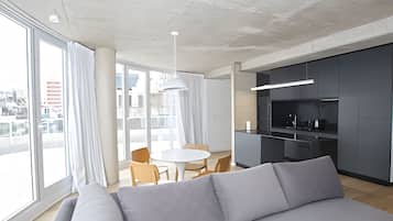 Un saló modern amb un sofà gris, una taula de dinar amb cadires i una zona de cuina amb armaris foscos i un taulell blanc.