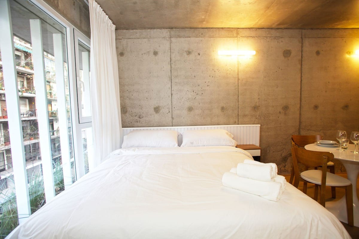 A hotel room with a large bed, a dining table with chairs, and a view of a building through a window.