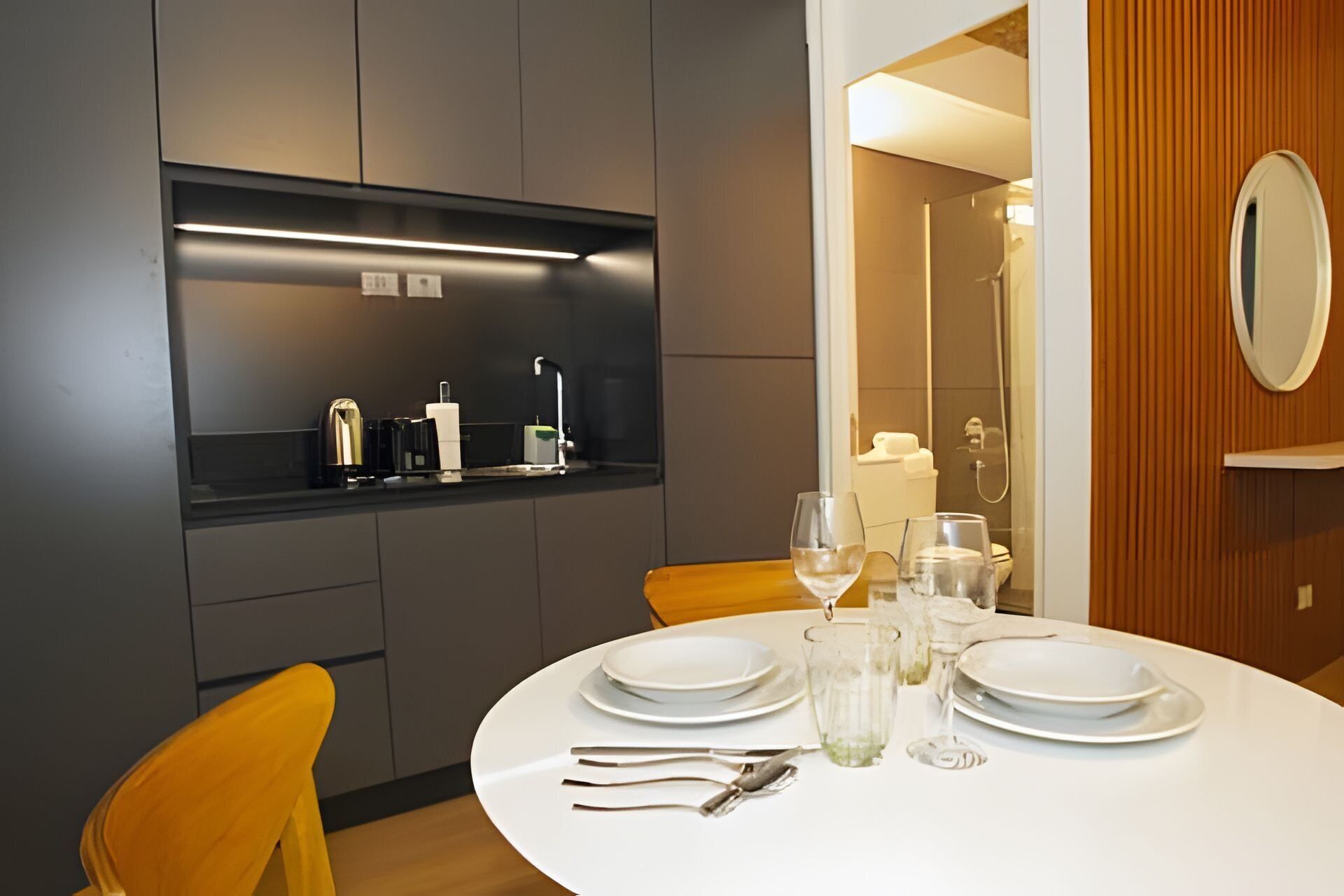 Een moderne eethoek met een ronde tafel gedekt voor vier personen, compleet met witte borden, glazen en bestek. Een inbouwkast boven de tafel bevat een koffieapparaat en andere voorzieningen.