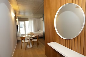 Habitación de hotel con espejo redondo, paneles de madera, una cama, una mesita con sillas y vistas al exterior.
