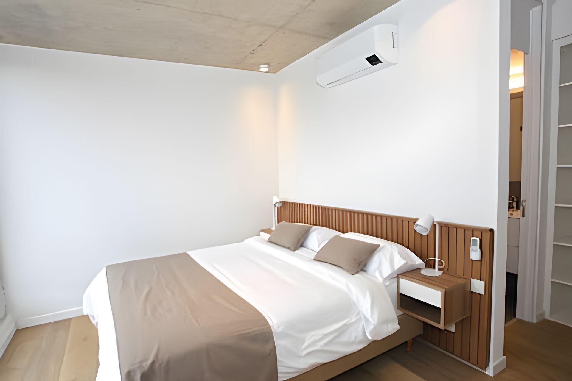 Un dormitorio moderno con cabecera de madera, mesita de noche con lámpara y aire acondicionado integrado.