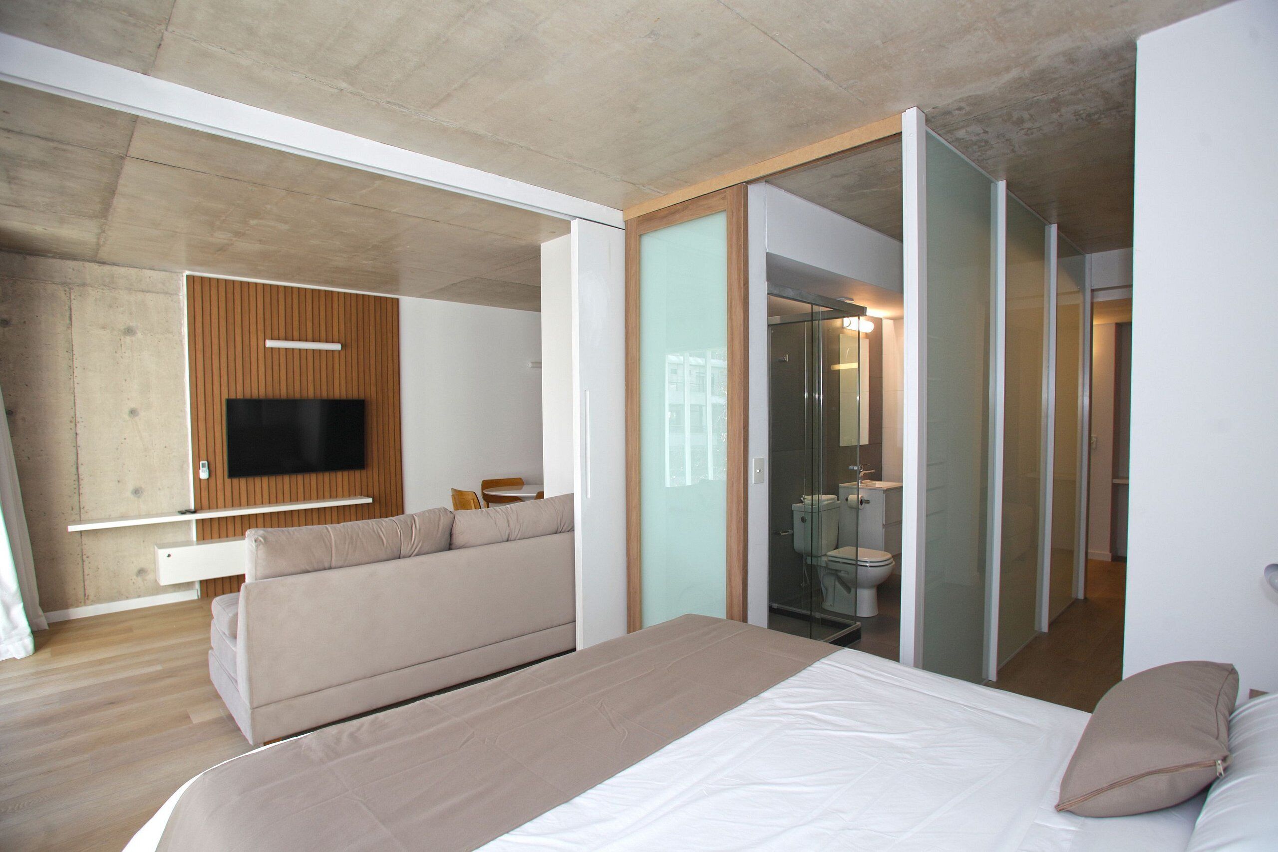 Una habitación de hotel moderna con una cama grande, un sofá, un televisor montado en un panel de madera y un baño con paredes de vidrio.