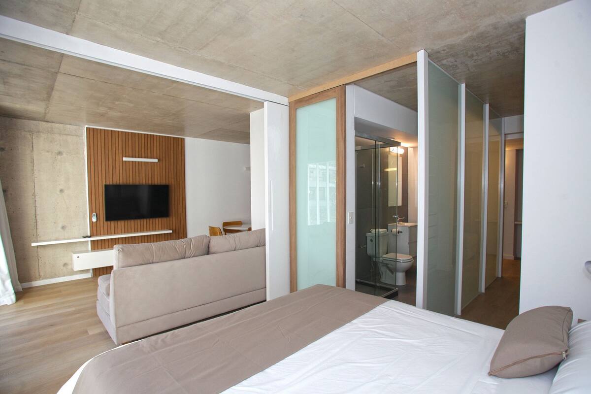 Una habitación de hotel moderna con una cama grande, un sofá, un televisor montado en un panel de madera y un baño con paredes de vidrio.
