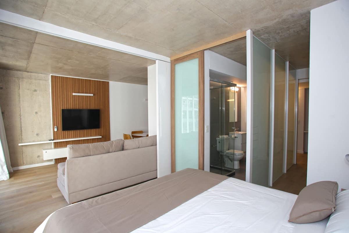 A modern hotel room with a large bed, a sofa, a TV mounted on a wooden panel, and a glass-enclosed bathroom.