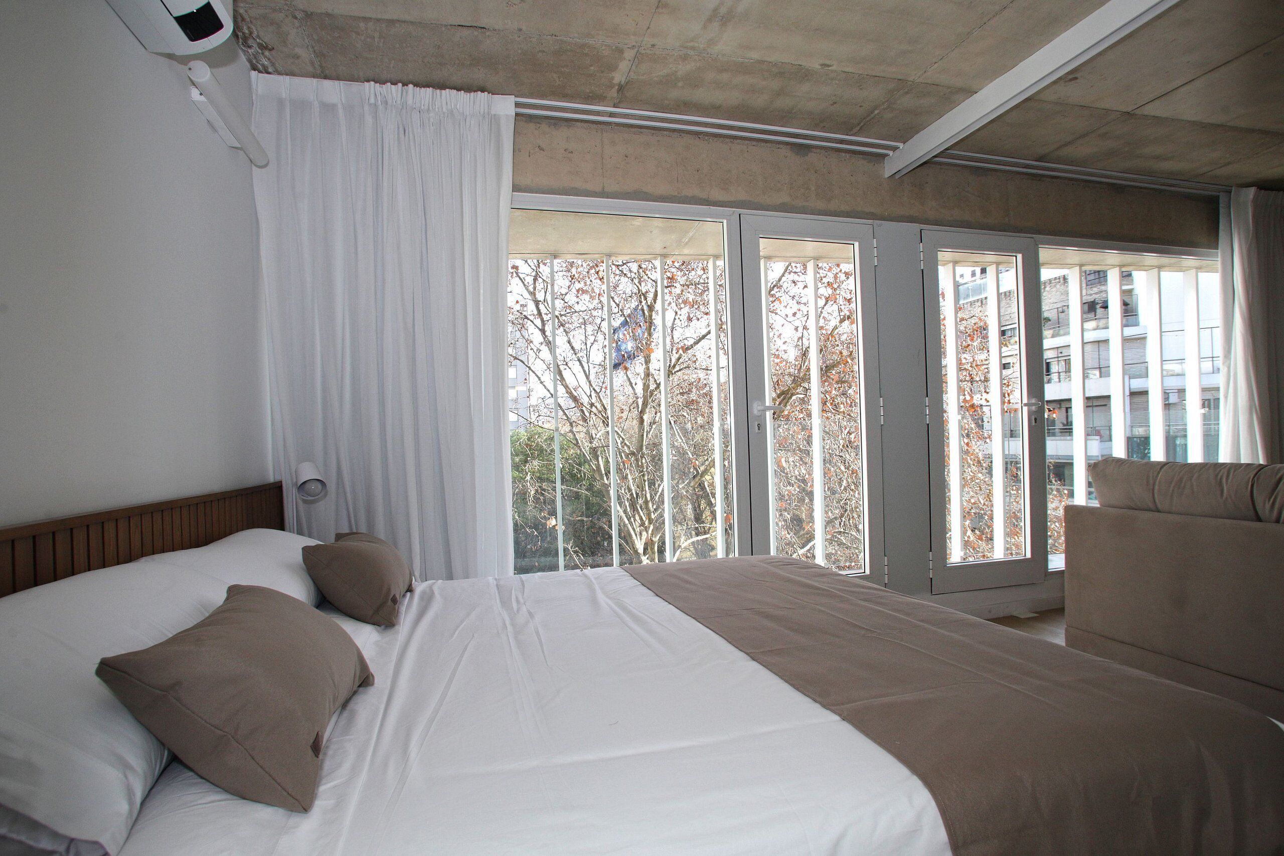 Quarto de hotel com uma cama grande, um sofá e uma porta de vidro deslizante que dá para uma varanda com vista para edifícios e árvores.