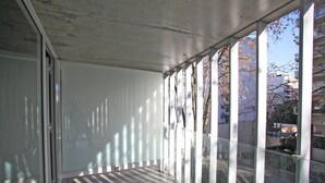 Un balcón con amplias ventanas, techo de hormigón y vistas a edificios y árboles.