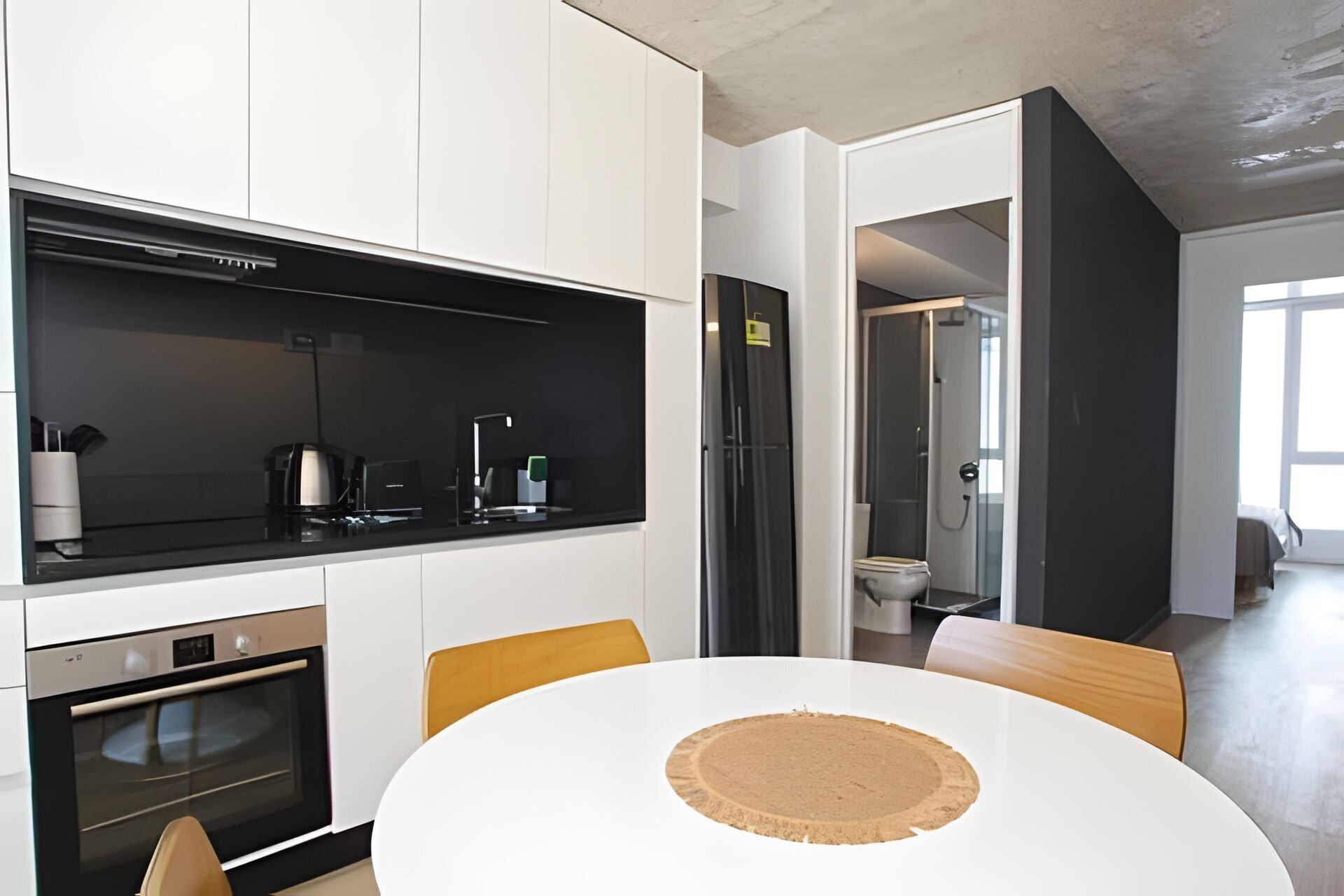 A modern kitchen with white cabinets, a black countertop, and a built-in oven. There is a round table with a cork mat in the center.