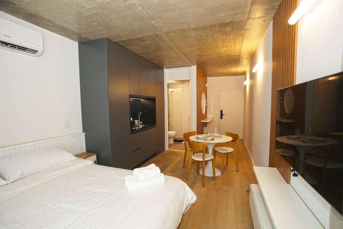 A hotel room with a bed, a TV, a dining table with chairs, and a small kitchen area.