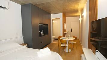 A modern hotel room with a bed, a built-in TV, a small dining area, and a shower.
