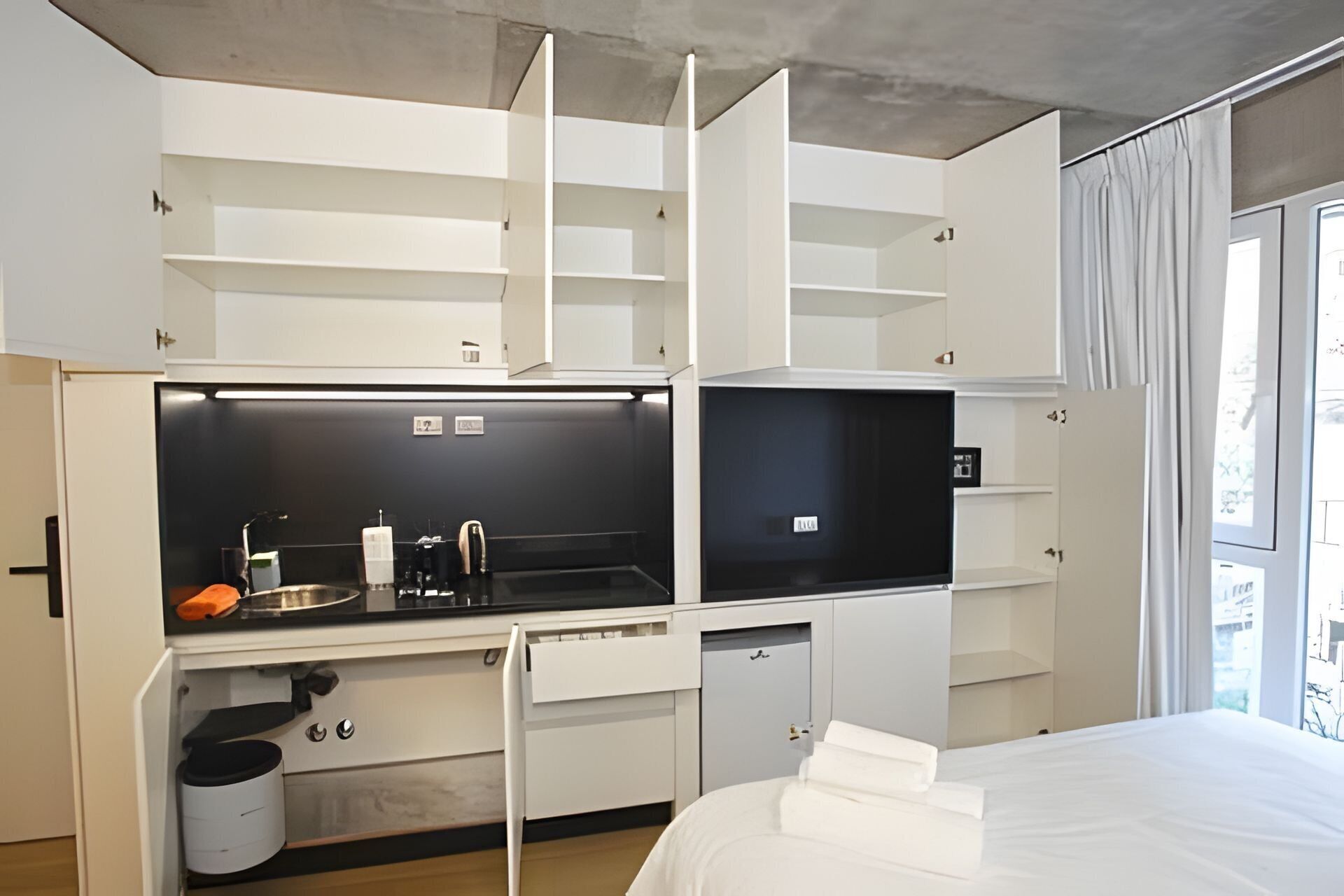 A compact hotel room with a kitchenette, a bed, and a window with curtains.