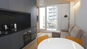 Cocina moderna con armarios grises, encimera negra y horno empotrado. Hay un comedor con mesa blanca y sillas amarillas. Un sofá y una lámpara de pie están en la sala de estar.