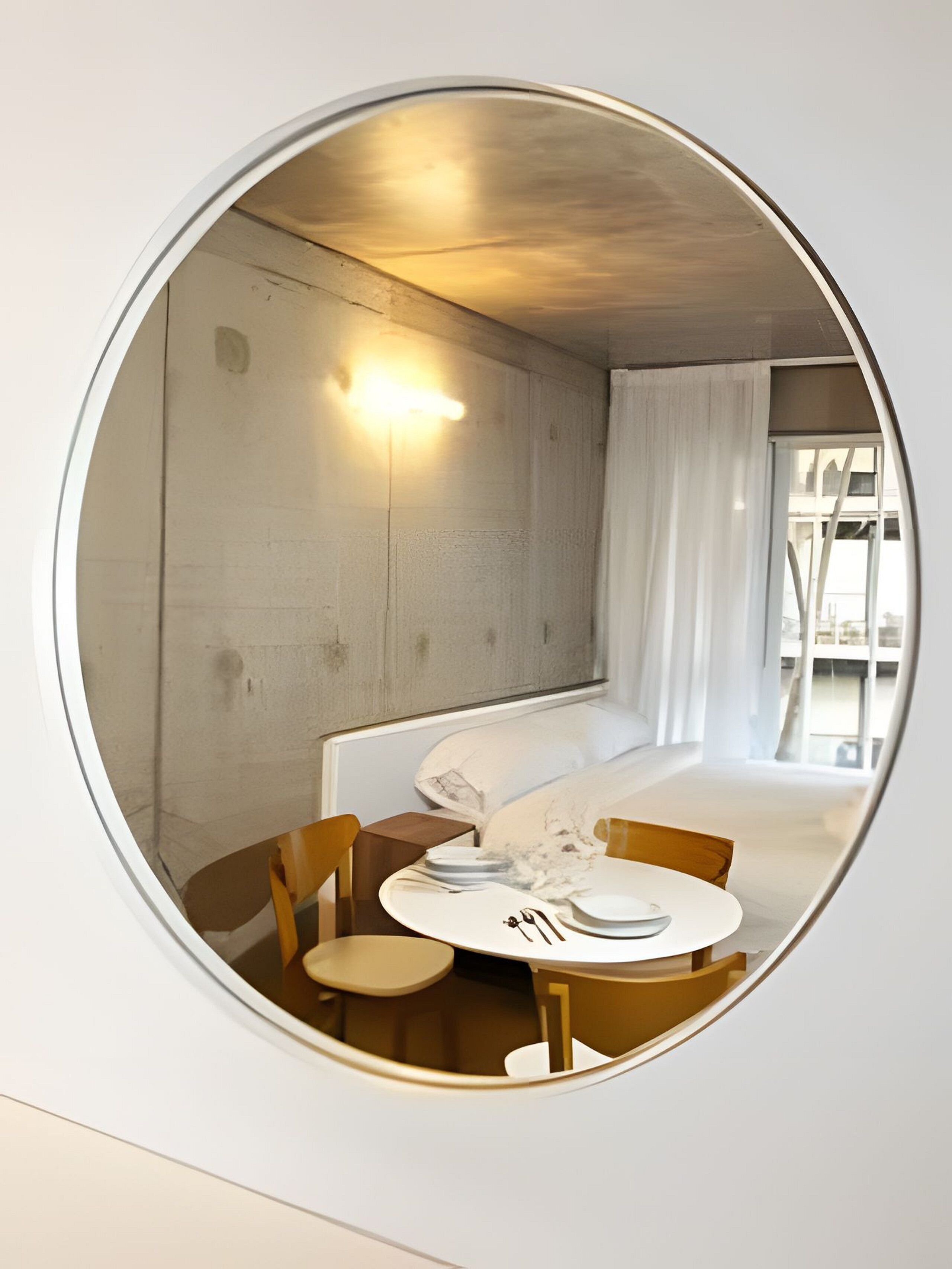Un espejo circular refleja una habitación moderna con una cama, una mesa con sillas y una lámpara fijada a la pared.