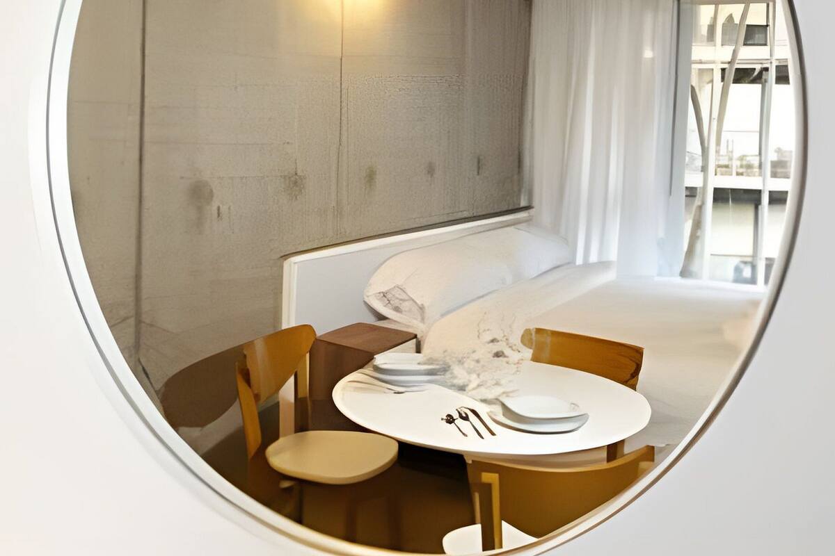 Un espejo circular refleja una habitación moderna con una cama, una mesa con sillas y una lámpara fijada a la pared.