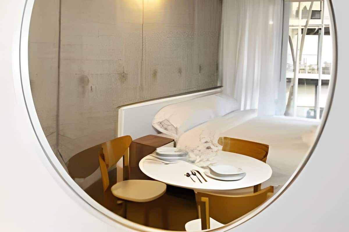 A circular mirror reflecting a modern room with a bed, a table with chairs, and a wall-mounted light.