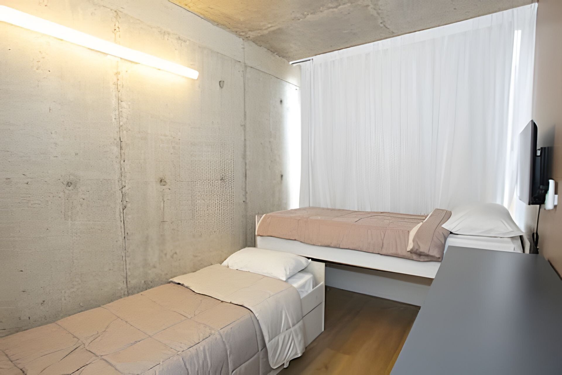 A small, compact room with a bunk bed and a single bed, a flat-screen TV, and a window with sheer curtains.